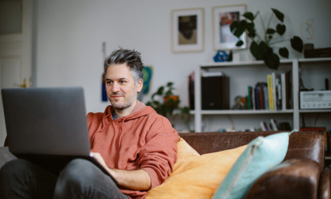 Ein Mann sitzt in seiner Wohnung auf dem Sofa und hat seinen Laptop auf dem Schoß.