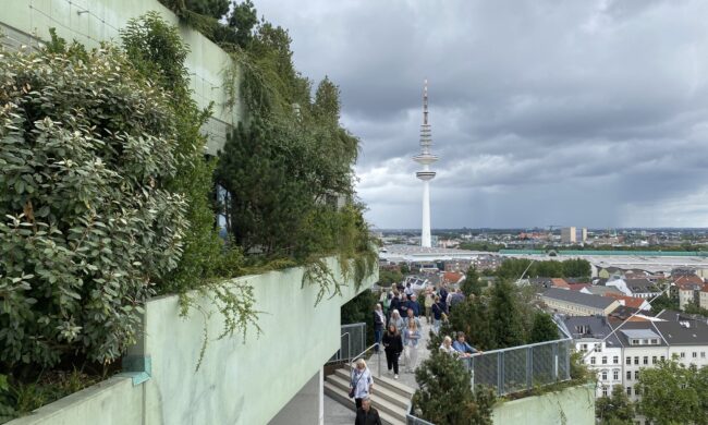 Menschen besuchenden Grünen Bunker in Hamburg, umgeben von Pflanzen und Bäumen, mit Blick auf den Hamburger Fernsehturm