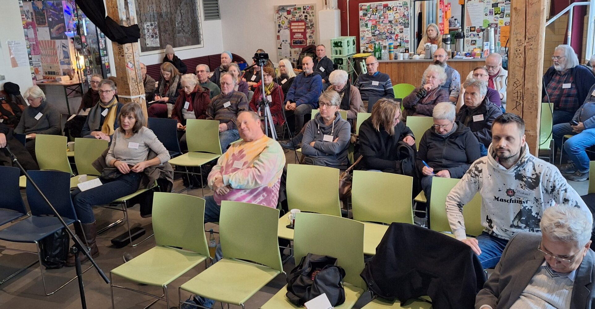 Eine Versammlung der alternativen Genossenschaftskongress. Mehrere Menschen sitzen in einem Raum auf Stühlen und interagieren miteinander.