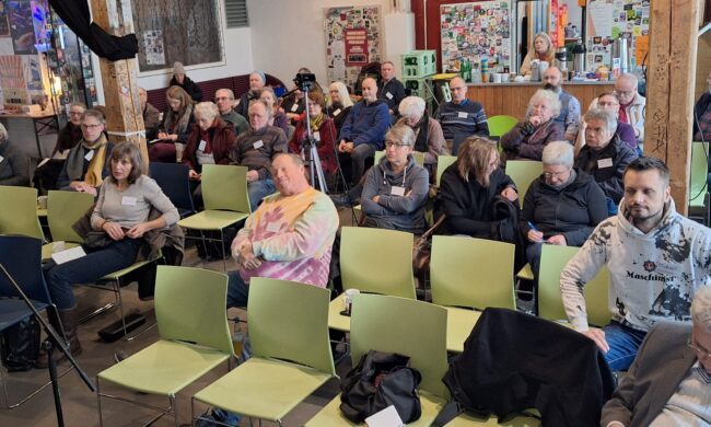 Eine Versammlung der alternativen Genossenschaftskongress. Mehrere Menschen sitzen in einem Raum auf Stühlen und interagieren miteinander.